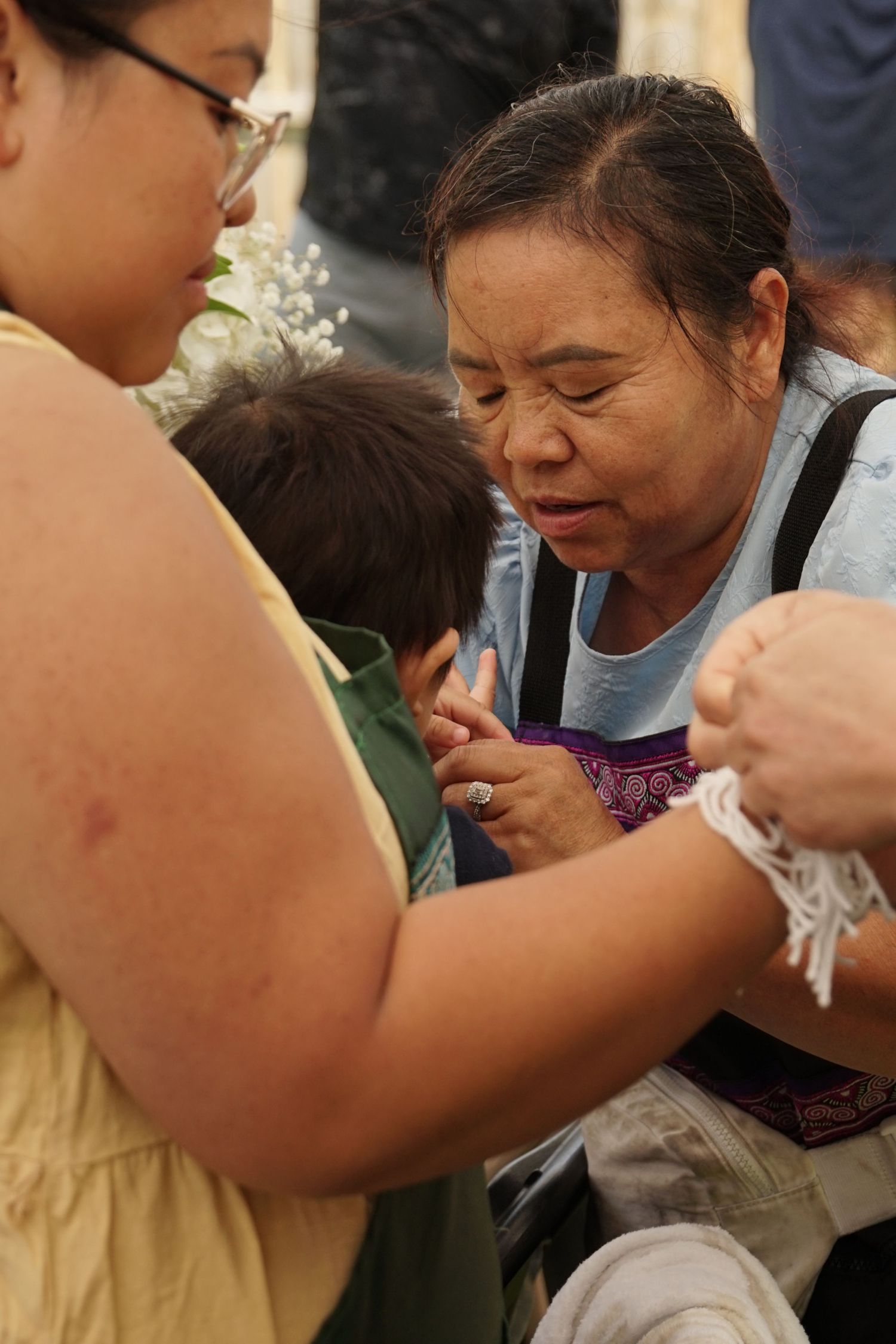 Elderly Asian woman in blue shirt consoles child while another woman looks on in an emotional outdoor gathering.