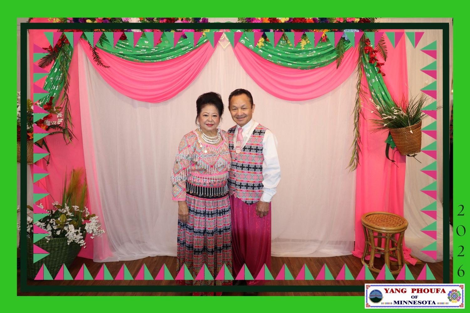 Couple posing together in traditional Hmong attire against a decorated pink and green draped backdrop at a formal event.