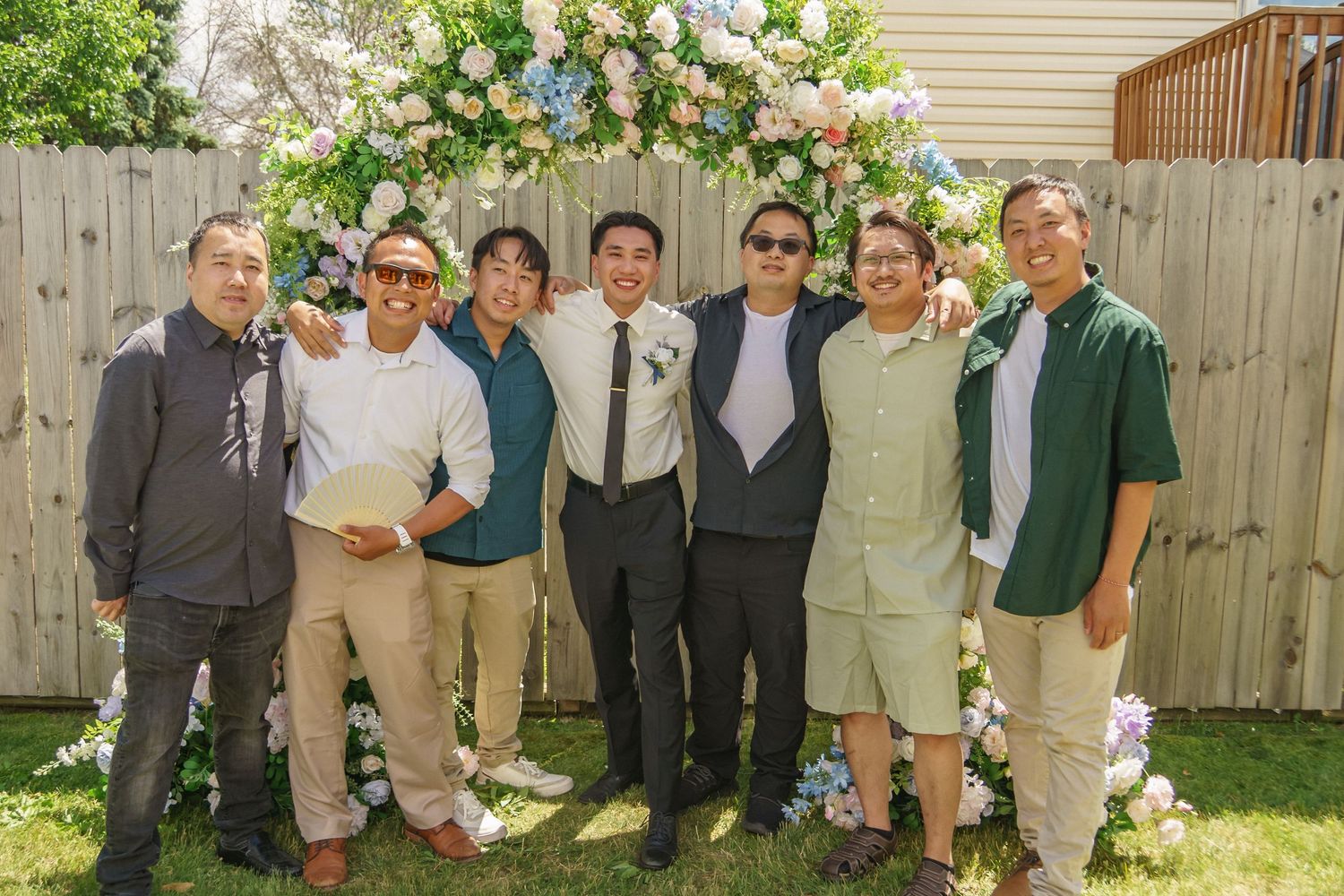 Seven men smile together in front of a colorful floral arch at a sunny outdoor wedding reception.