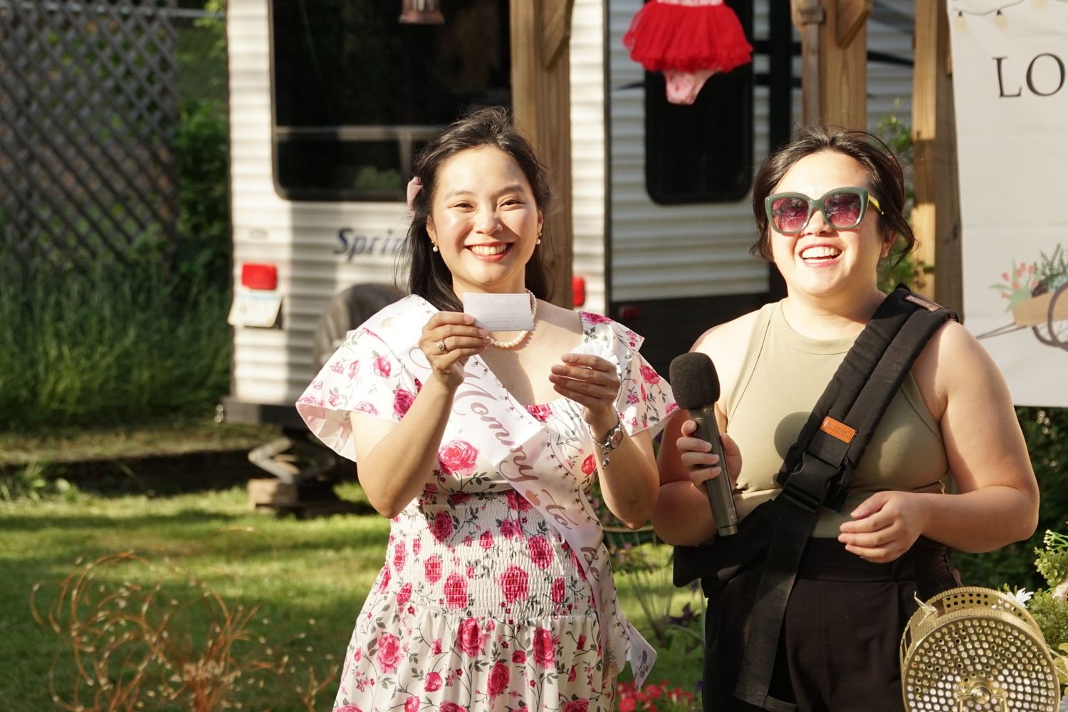 Two smiling Asian women outdoors, one pregnant in floral dress, other carrying baby in carrier near trailer.