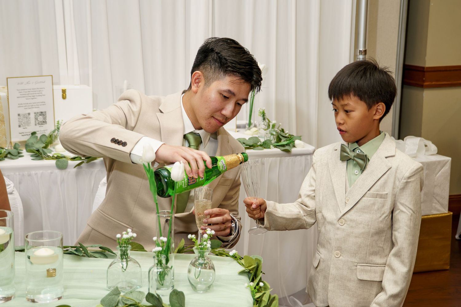 Man in beige suit pouring champagne for a boy in matching suit at an elegant wedding reception.