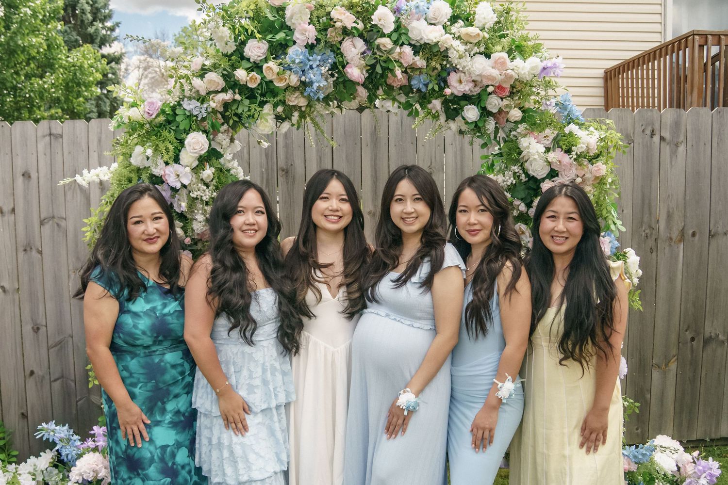 Six young women smiling together under a pink and blue floral arch at an outdoor party.