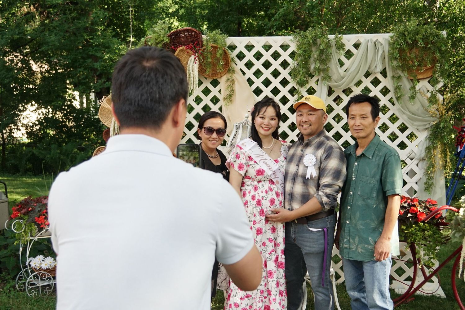 A photographer captures a group of five people posing outdoors near a decorative white lattice backdrop at a garden event.