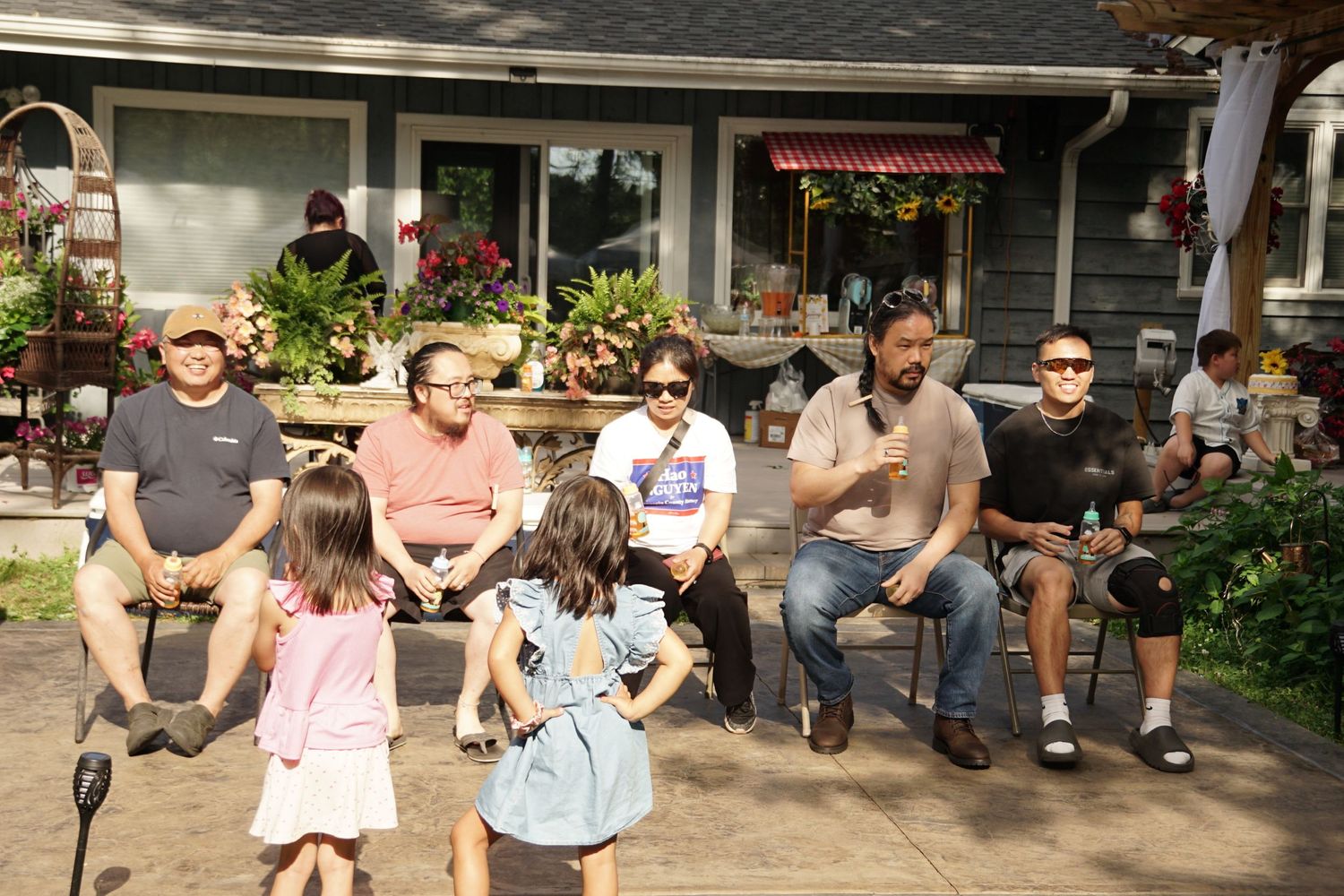 Family gathering outdoors on sunny day, adults seated on porch steps with children playing in foreground, surrounded by colorful flowers.