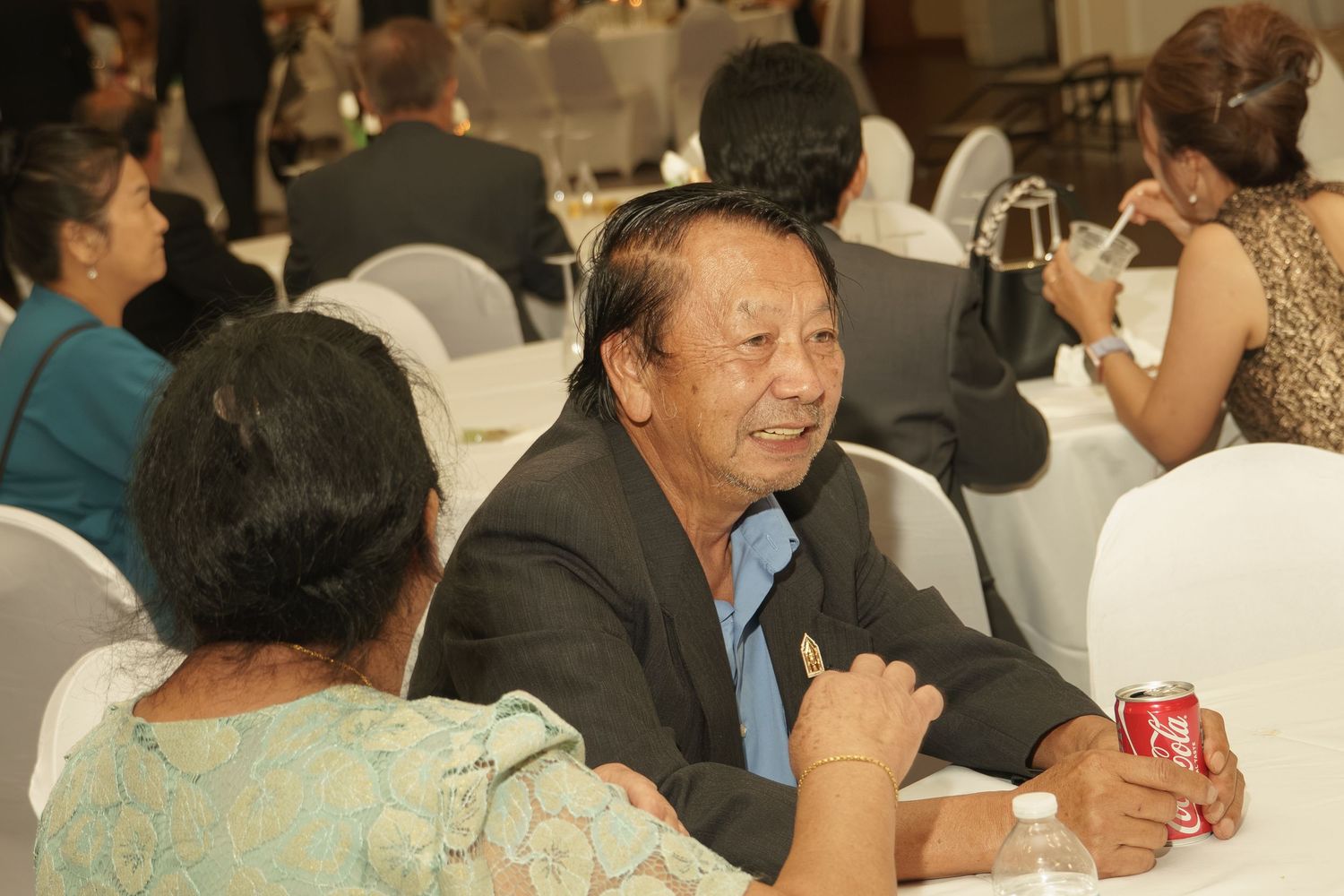 Elderly man smiling at a banquet table holding a Coca-Cola can, socializing with guests at a formal event.