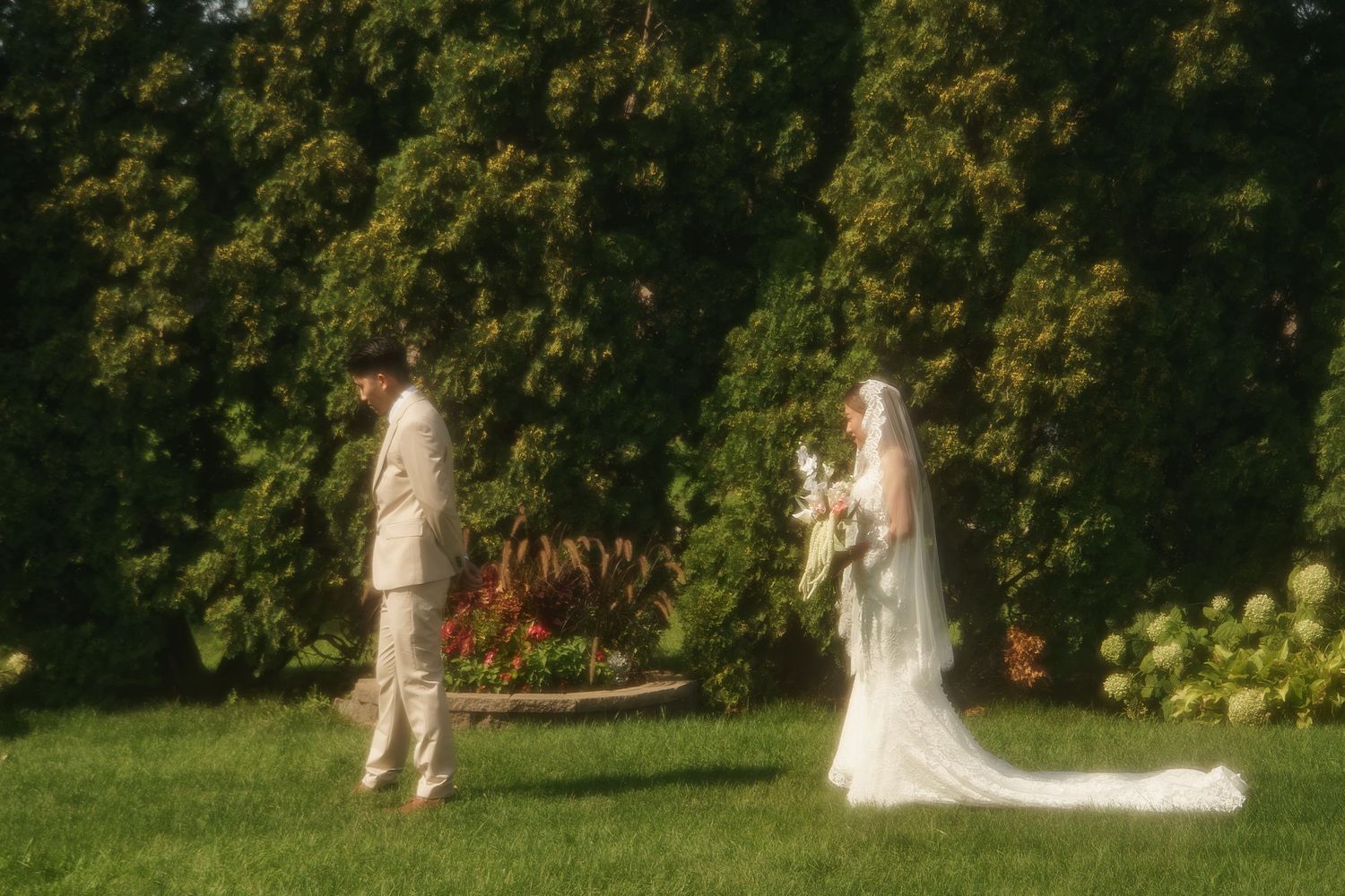 Bride and groom stand facing each other on lush green lawn surrounded by tall trees during outdoor wedding ceremony.