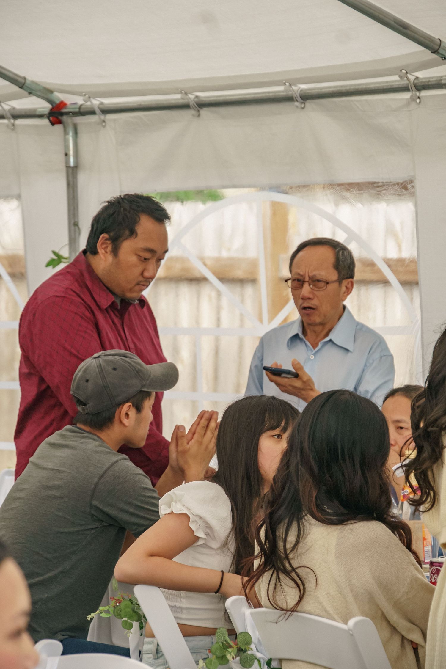 Men engage in conversation with seated guests under white tent at outdoor celebration.