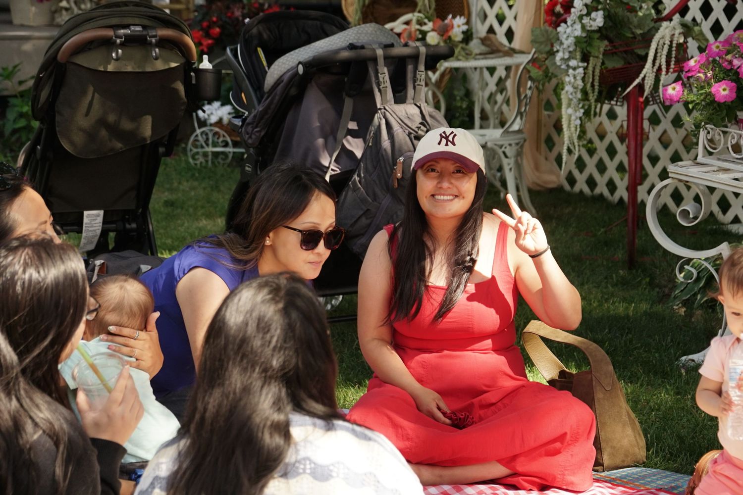 Women sitting on grass at an outdoor gathering, one in red outfit wearing a NY cap, another holding a baby.