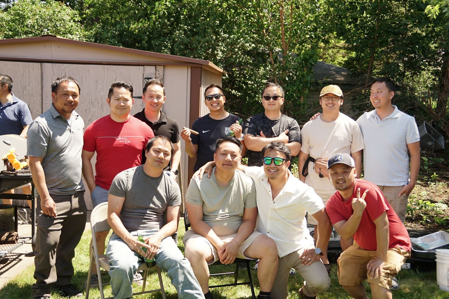 Large group of smiling men gathered outside near a shed on a sunny day.
