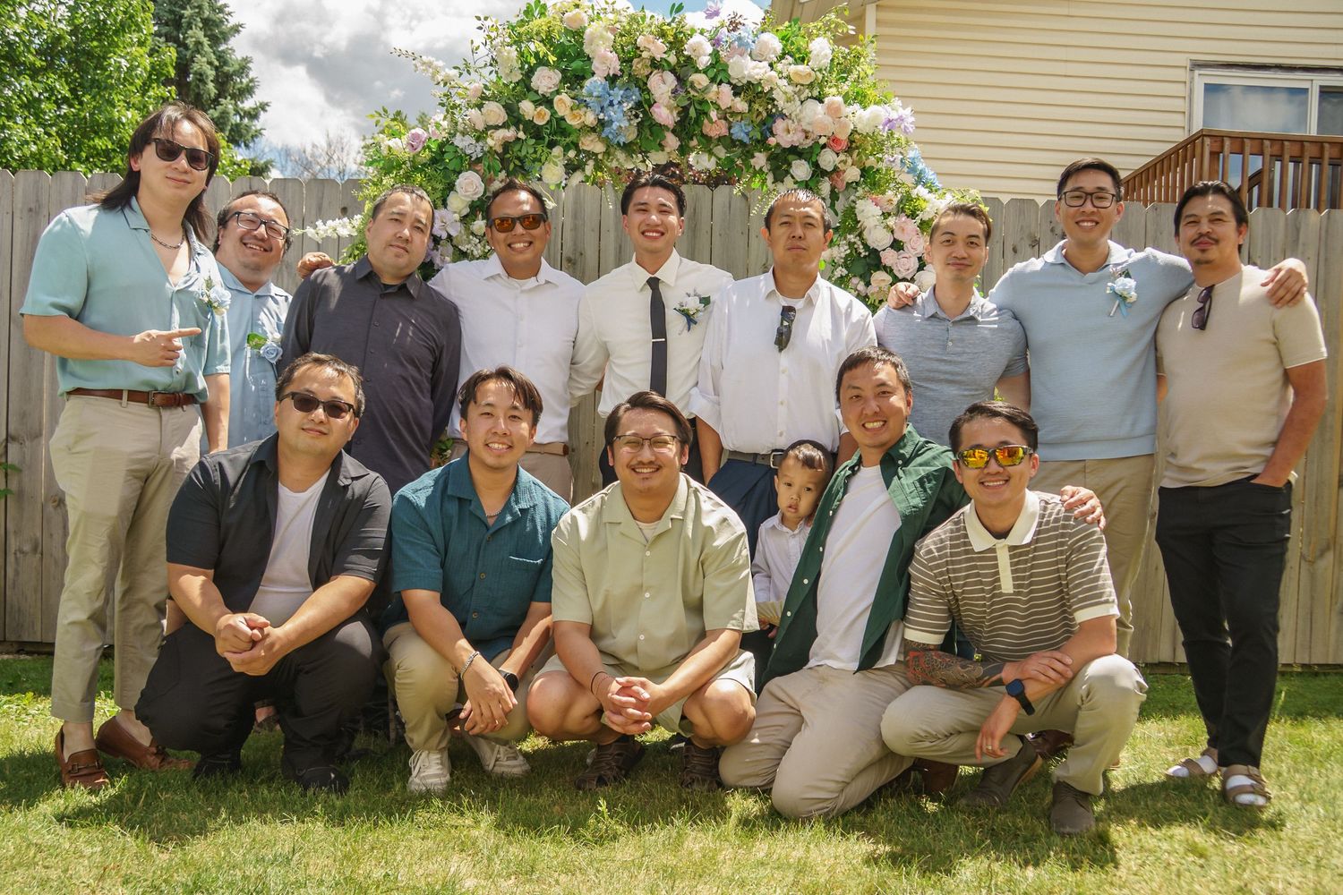 Large group of men pose together on green lawn in front of a floral arch at an outdoor wedding.