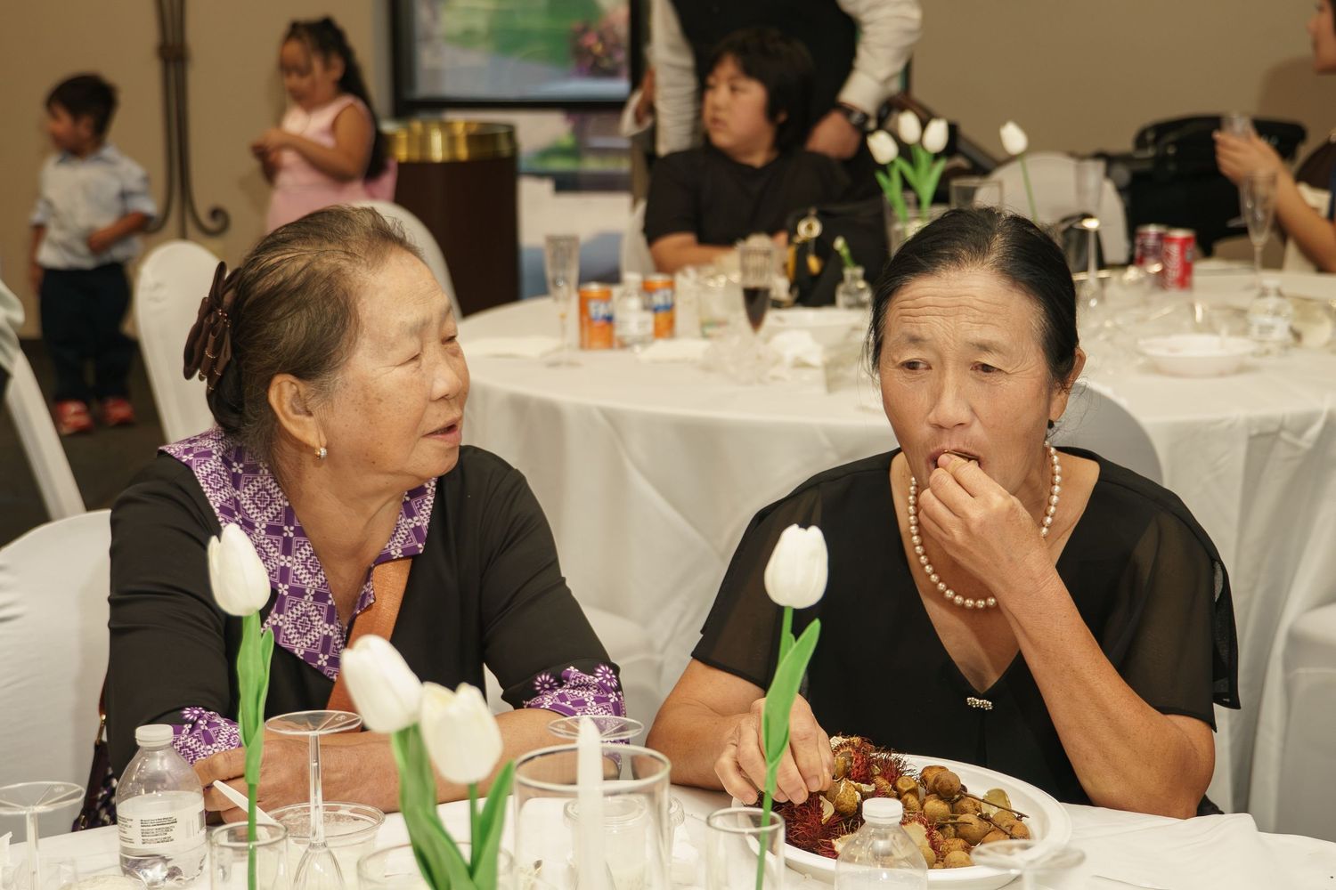 Two elderly Asian women in black attire converse at a banquet table adorned with white tulips.