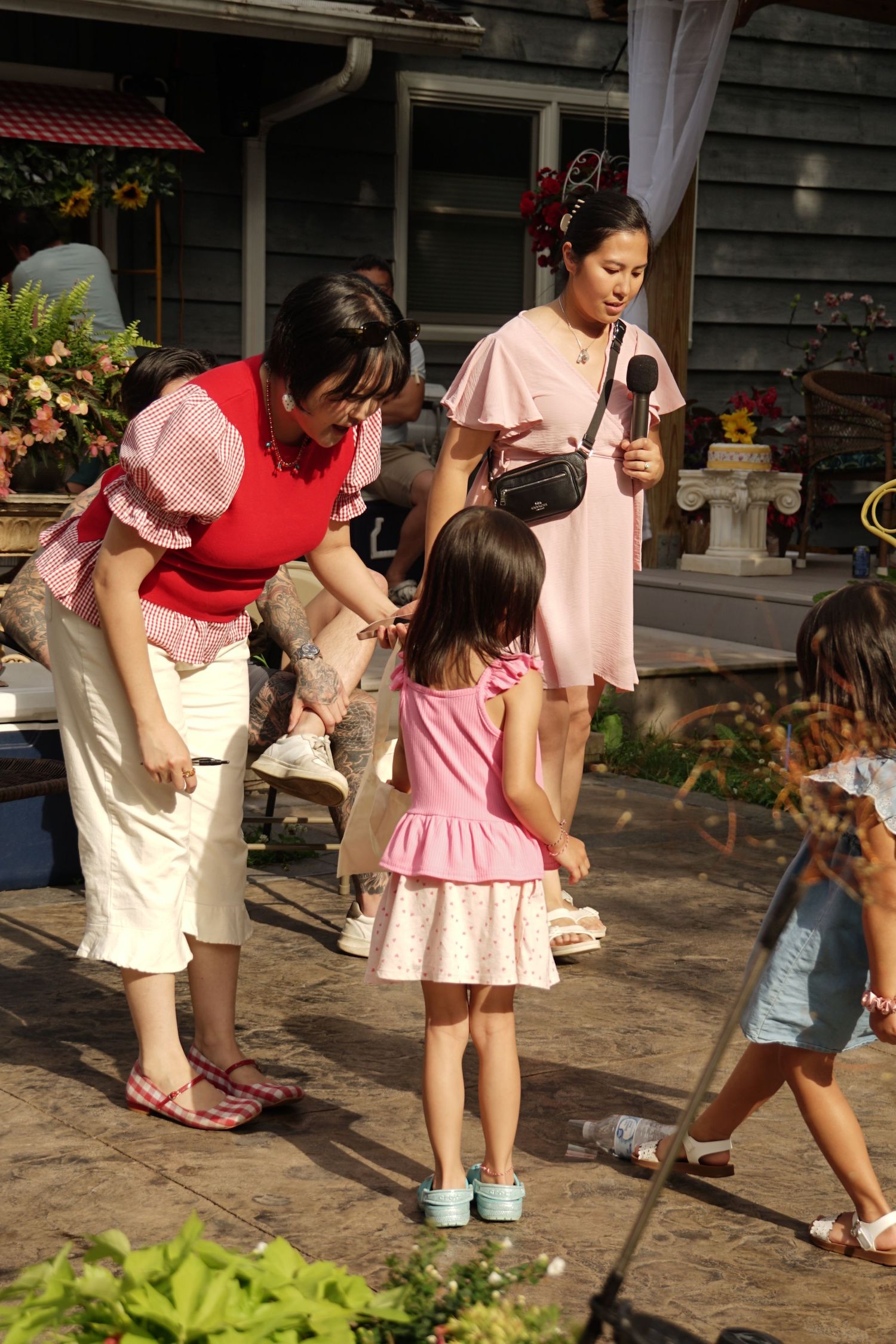 Two women in pink outfits interact with a young girl in a pink dress outdoors near a decorated wooden building.