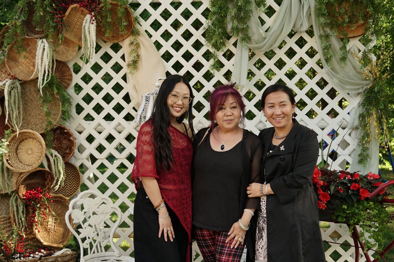 Three women smiling together in front of a white lattice backdrop decorated with greenery and woven baskets.