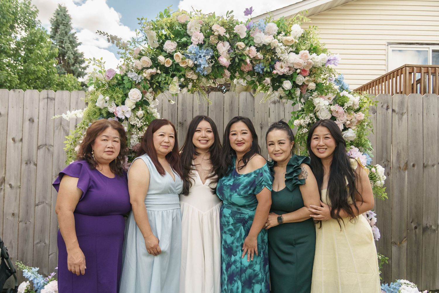 Six women posing together under a floral arch at an outdoor backyard celebration event.