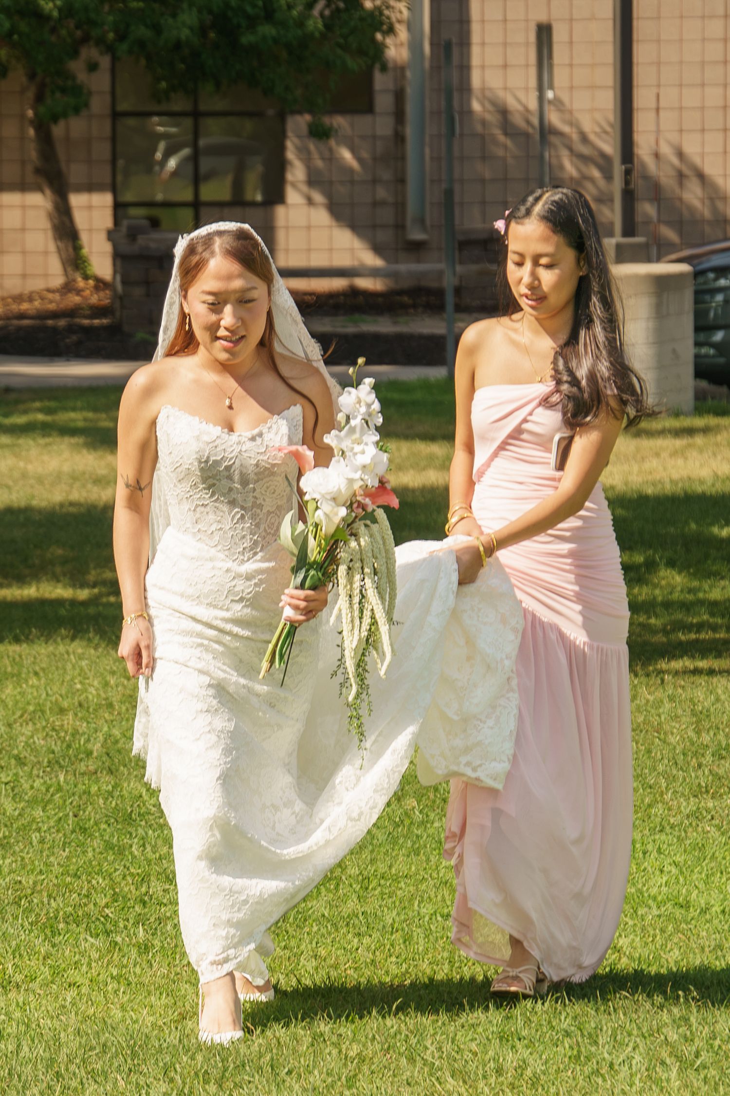 Bride in white lace gown holding bouquet walks on grass while bridesmaid in pink dress carries her train.