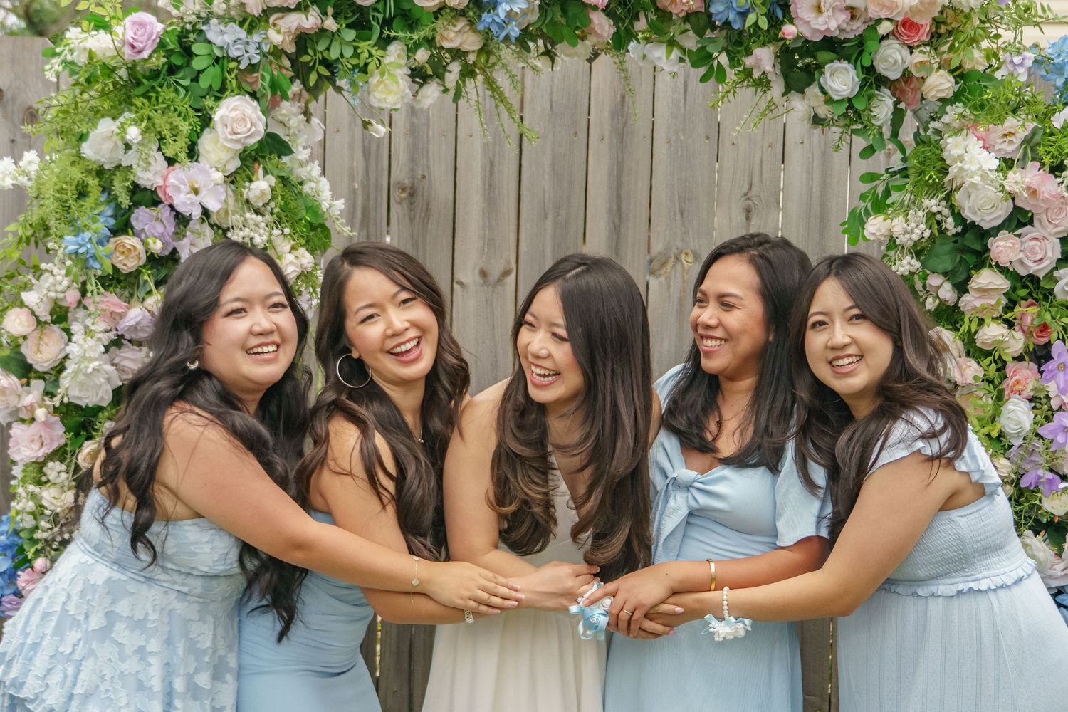 Five women in blue dresses laugh joyfully together under floral arch at baby shower.