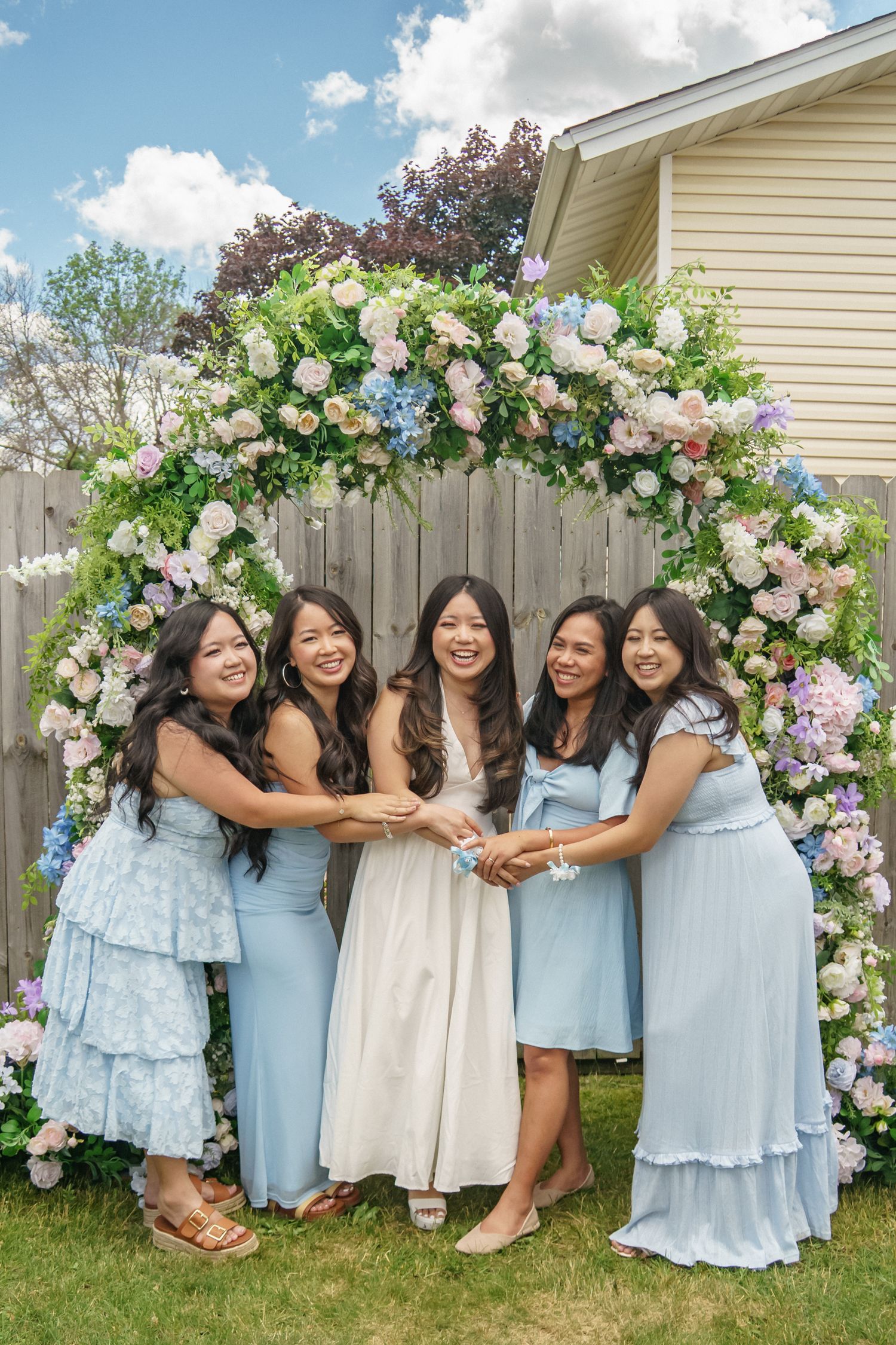 Bride in white dress with four bridesmaids in blue dresses under floral arch outdoors.