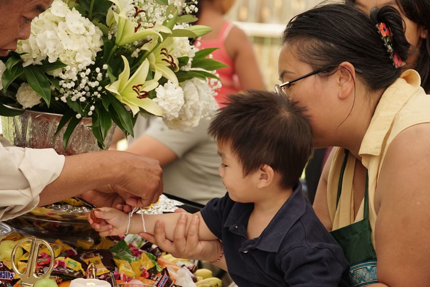 A woman and young child examine colorful items at a festive table adorned with white flowers at an Asian cultural celebration.