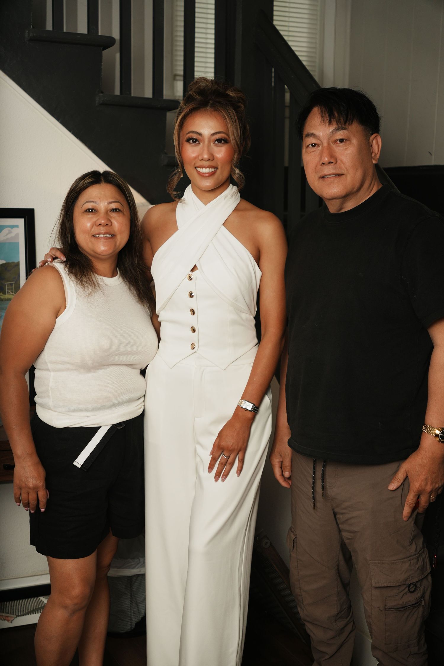 Three people posing together indoors, woman in white halter jumpsuit flanked by two others near a staircase.