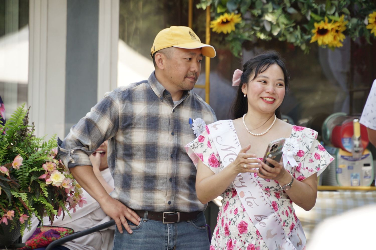 A man in a yellow cap and plaid shirt stands with a smiling woman in a floral dress holding cards outdoors.