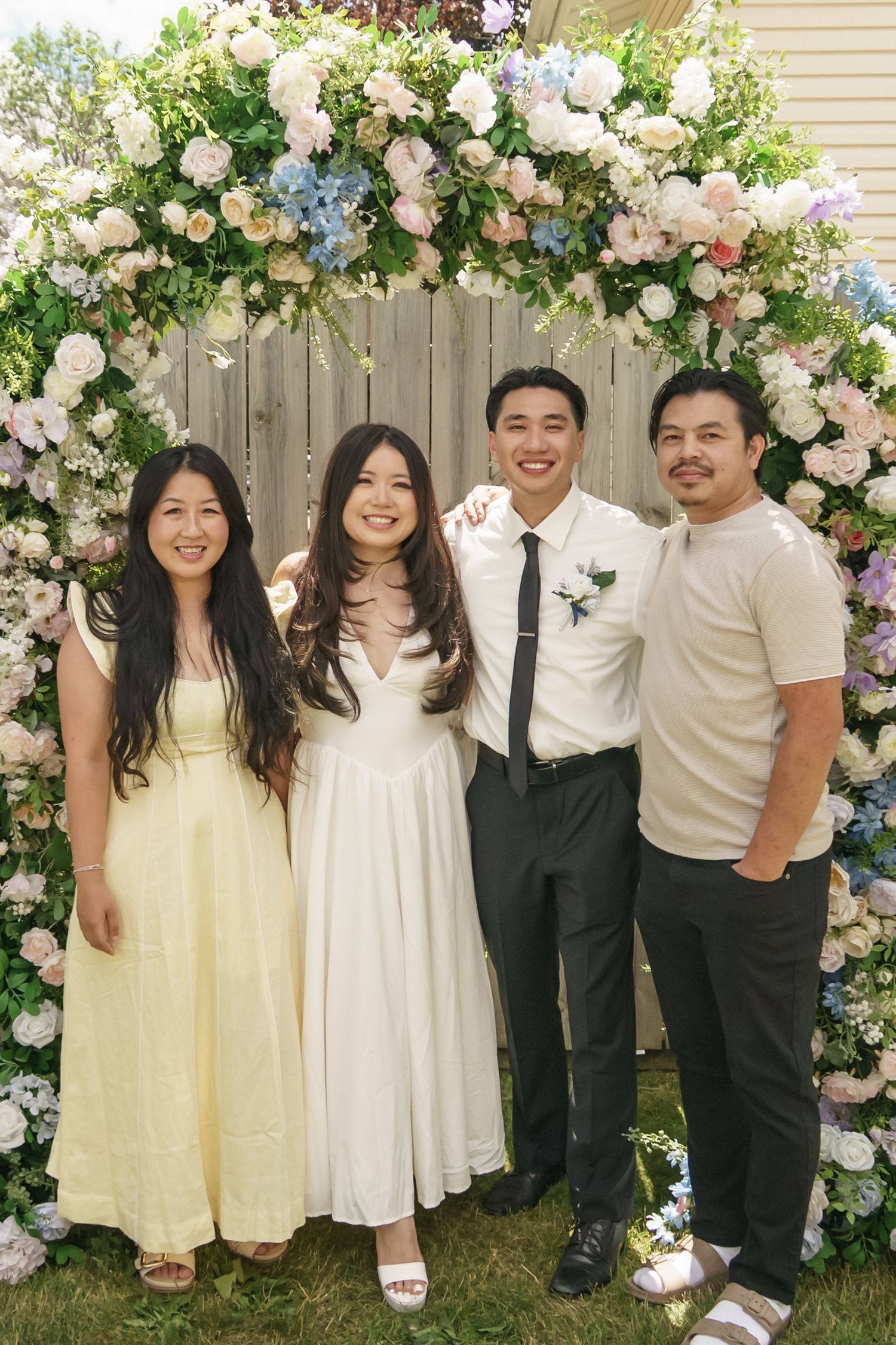 Bride in white dress and groom in suit pose with two guests under floral wedding arch.