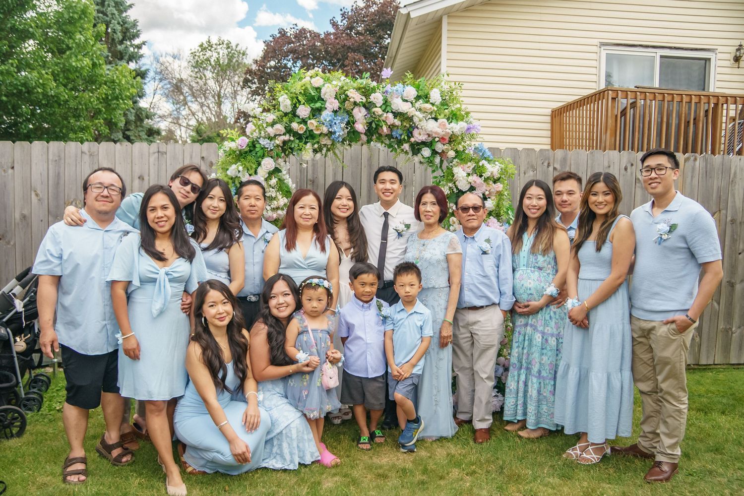 Large family group of about eighteen people posing together under a floral arch outdoors.
