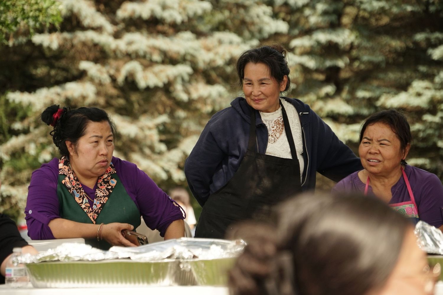 Three women smiling and chatting outdoors at a community gathering with food trays on a table.