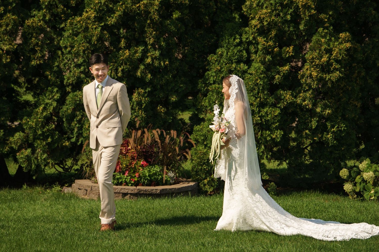 Bride in white lace gown with veil and groom in beige suit share first look moment on green lawn.