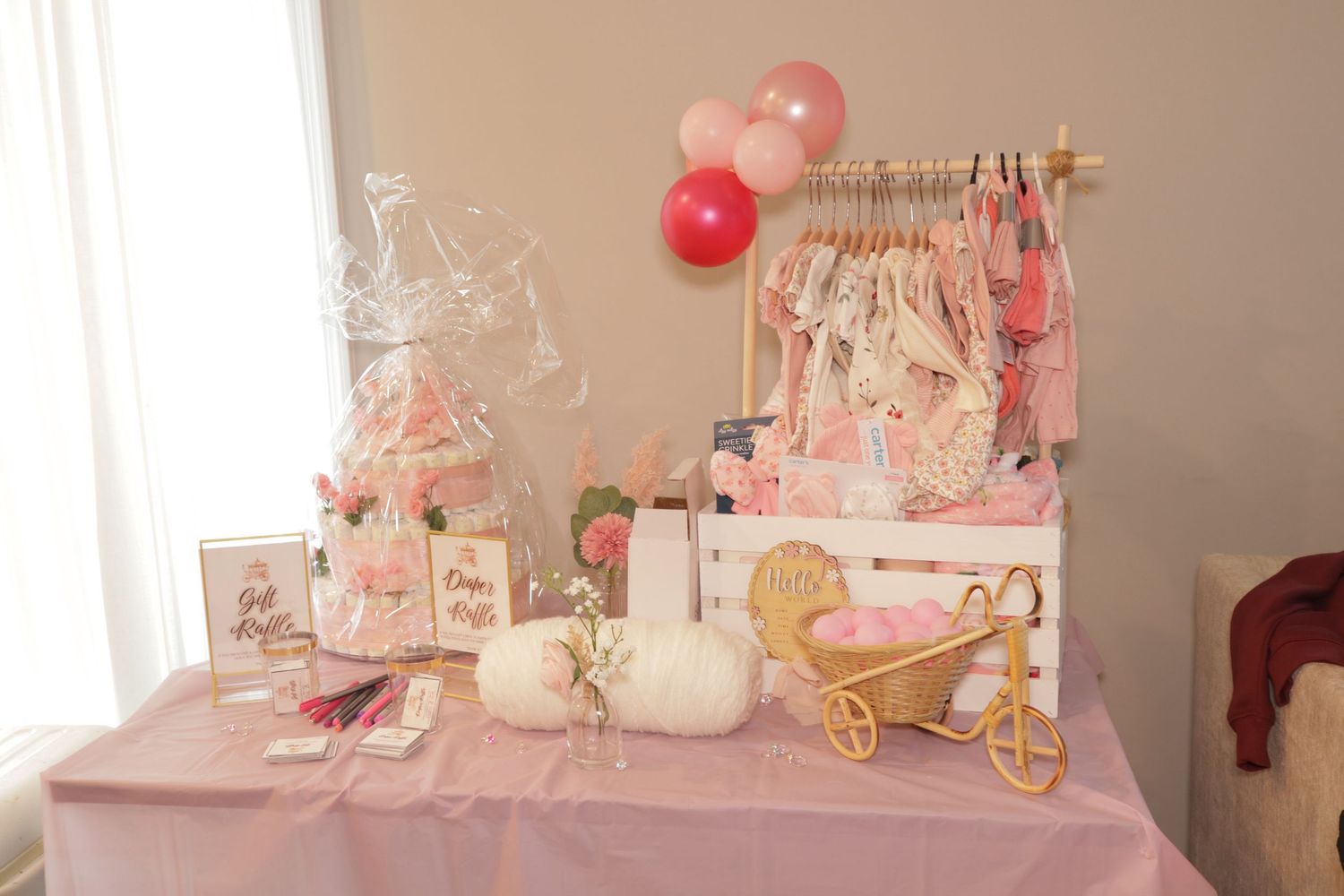 Pink baby shower gift table display with balloons, clothing rack, and decorative items.