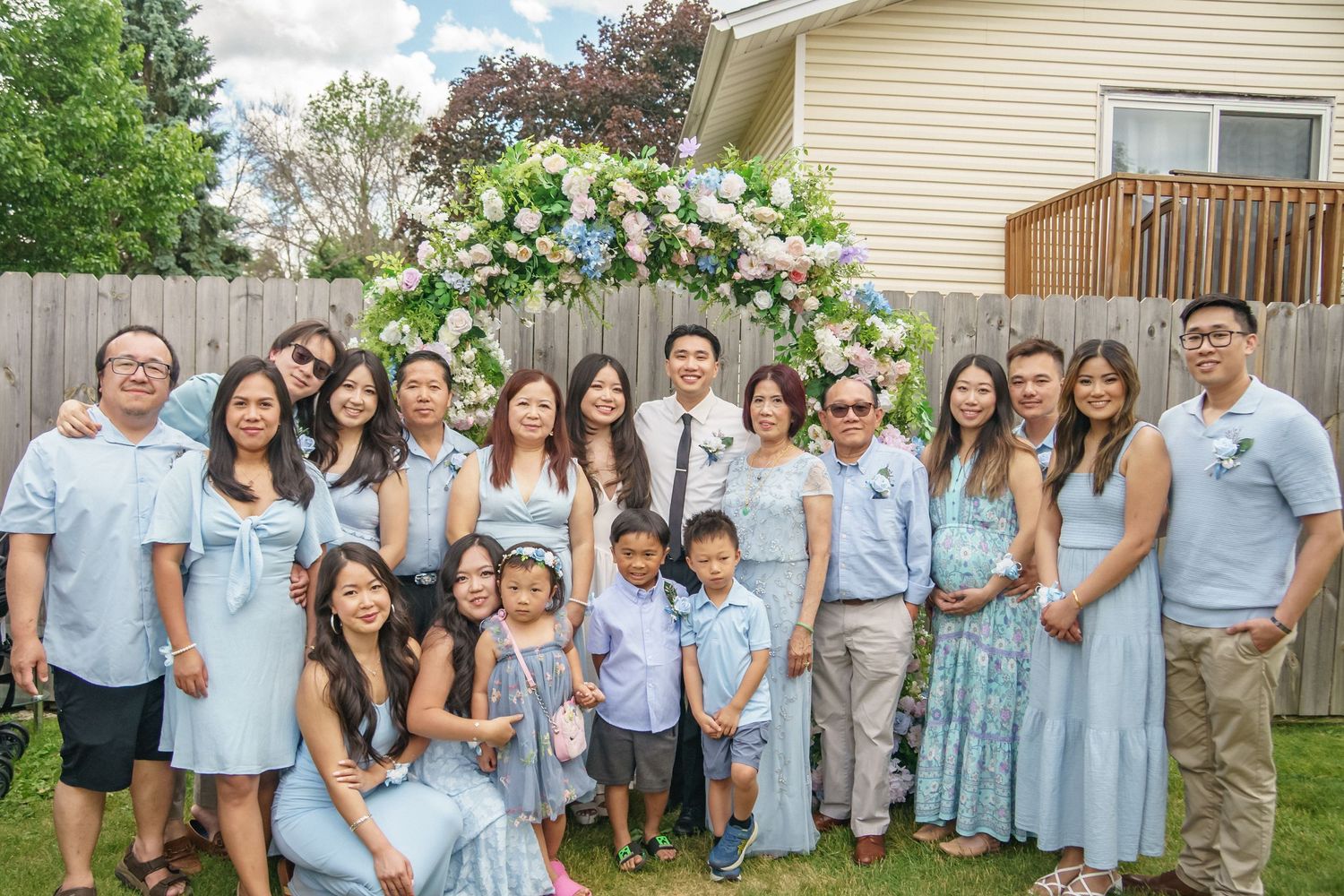 Large Asian family group photo at outdoor backyard wedding ceremony with floral arch backdrop.