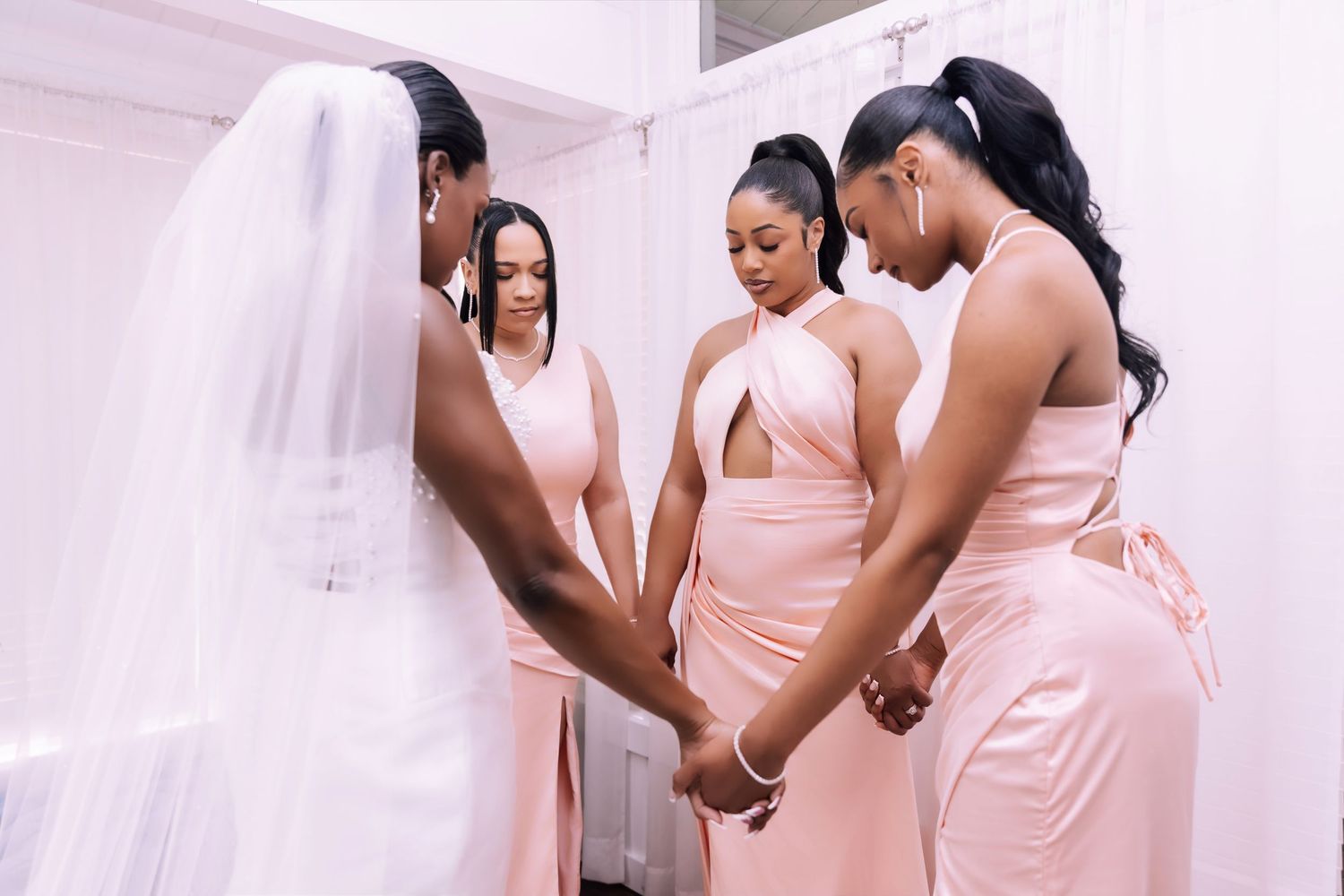 Bride and bridesmaids in blush dresses holding hands in prayer circle before wedding ceremony.