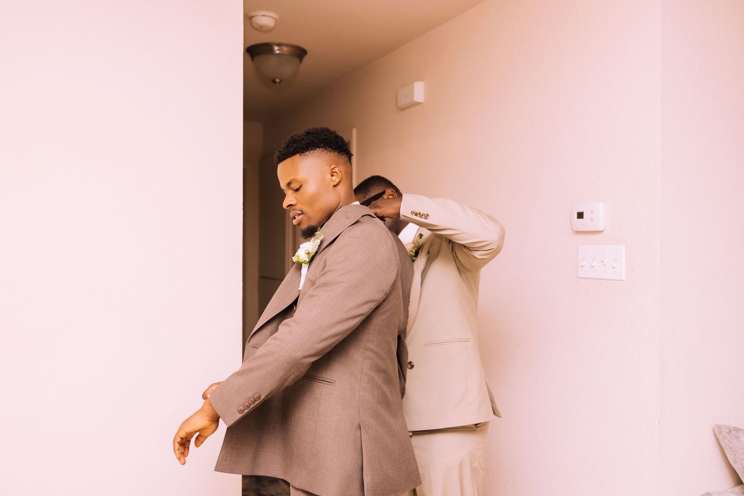 Groom in a brown suit with a boutonniere adjusts his sleeve while a groomsman in a tan suit fixes his jacket in a softly lit room.