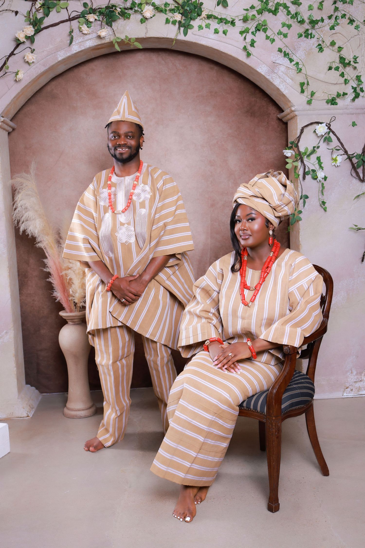 Couple in matching gold and white striped Nigerian Yoruba traditional attire with red coral beads posing for a portrait.