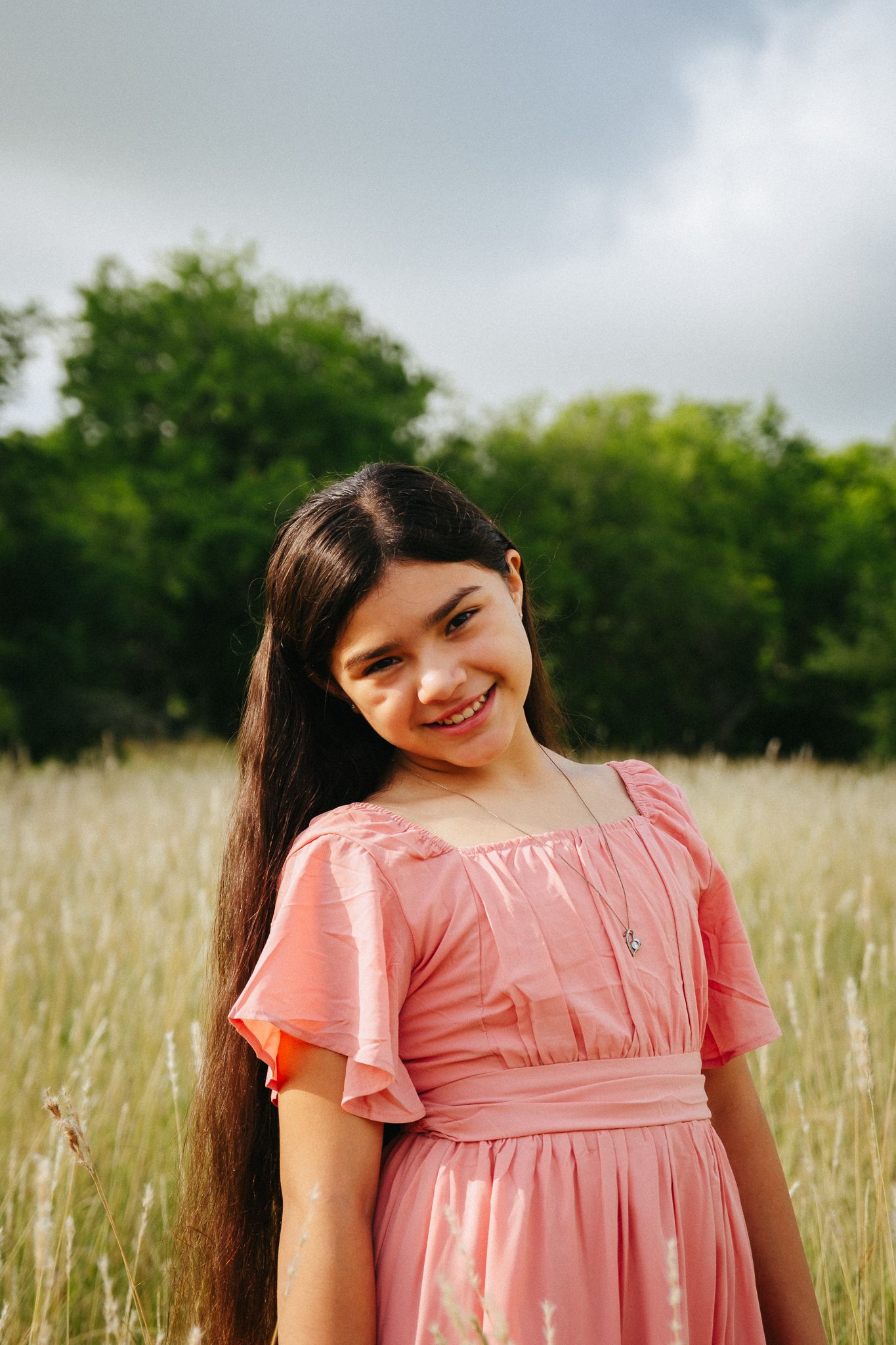 Young person in pink dress smiling while standing in tall grass field on cloudy day.