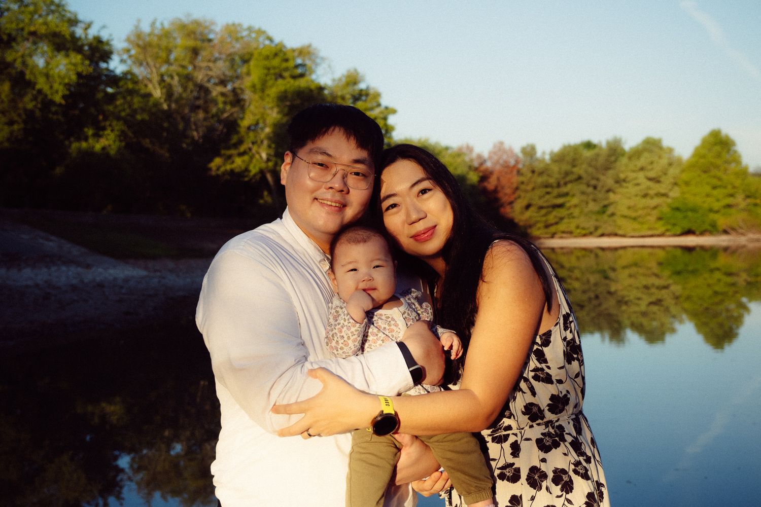 Romantic couple embraces outdoors by a serene lake with trees and greenery in the background.