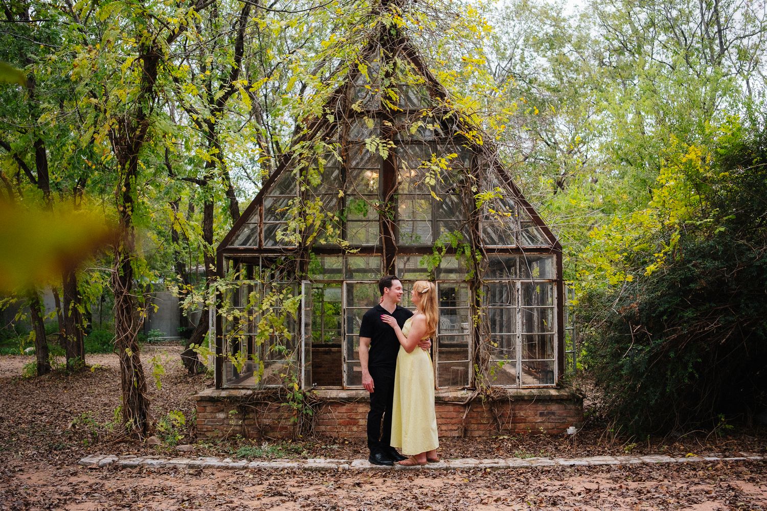 Couple embracing in front of a rustic glass greenhouse surrounded by lush green trees during an outdoor engagement session.