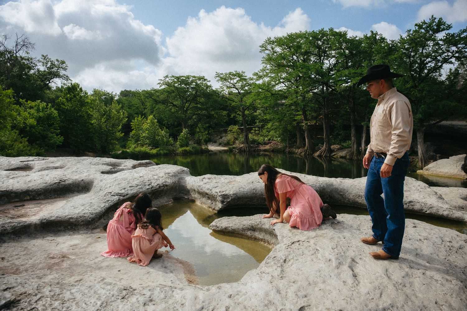 A cowboy watches two girls in pink dresses explore a natural rock pool surrounded by lush Texas Hill Country trees.