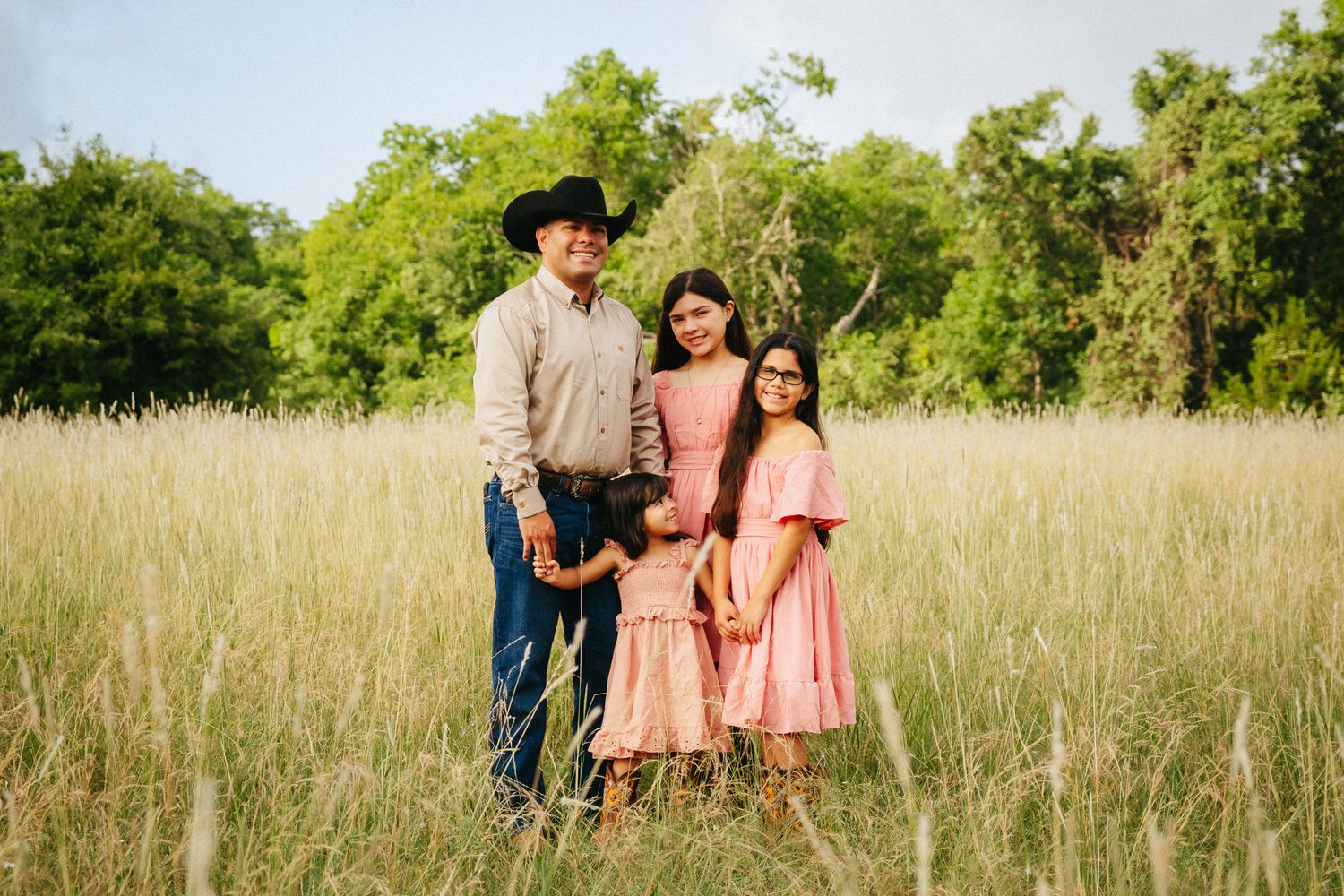 Family of four posing in golden meadow with lush green trees, wearing coordinating pink dresses and cowboy hat.