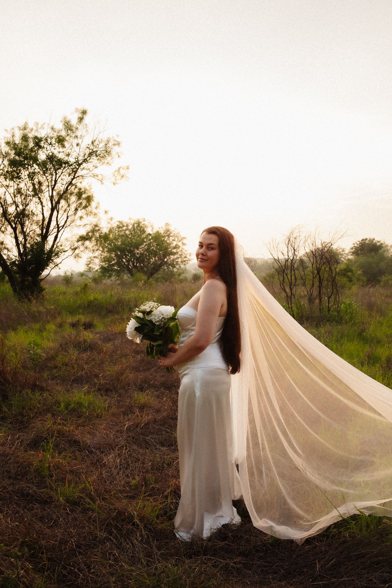 Bride in white gown and flowing veil holds bouquet in golden sunset field surrounded by trees.