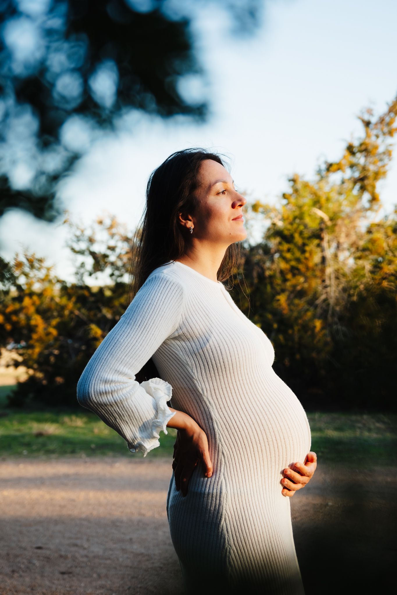 A pregnant woman in a white ribbed dress stands in nature during golden hour, cradling her baby bump.