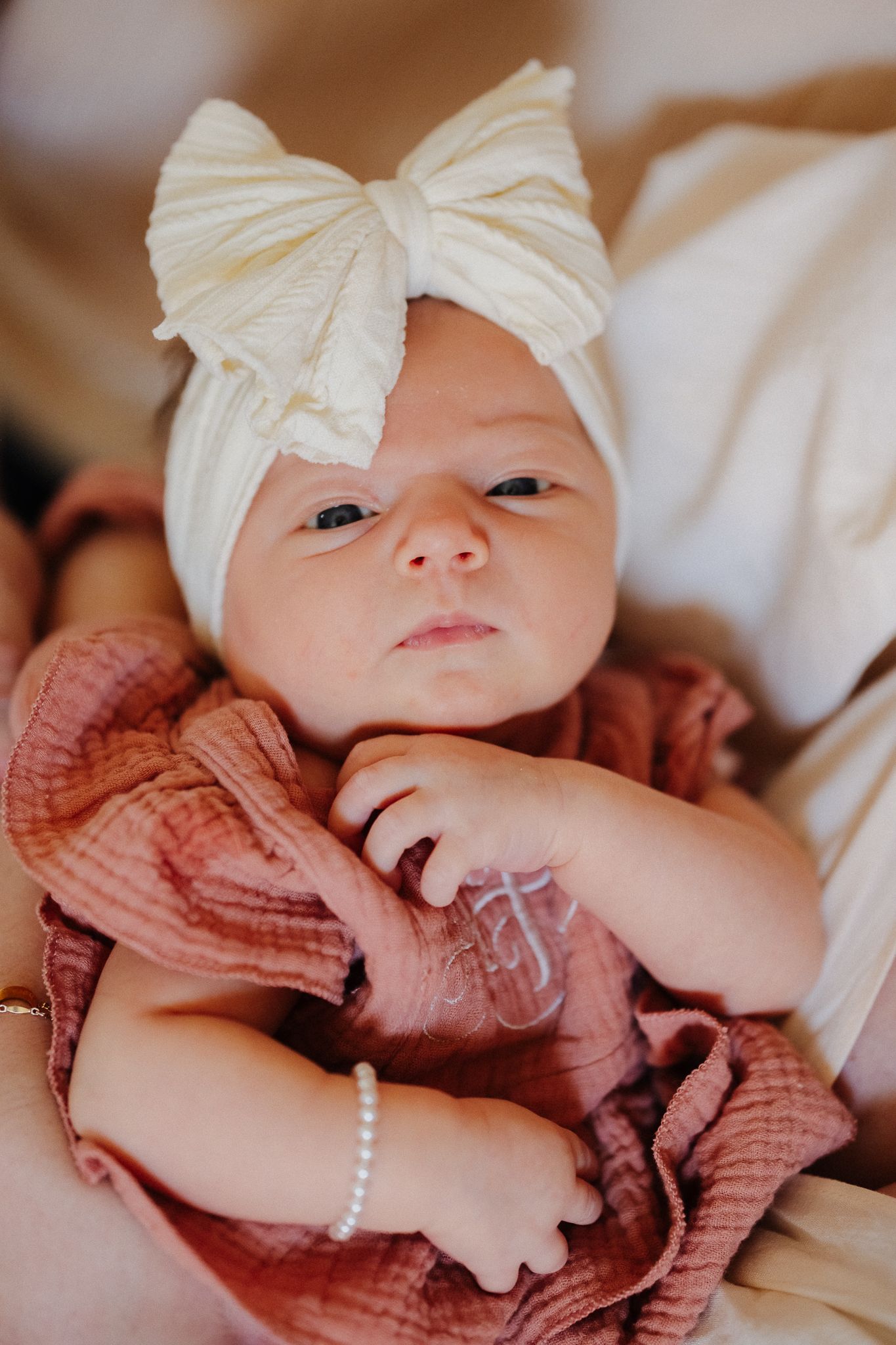 Newborn baby girl in pink ruffle outfit and cream bow headband awake and alert.