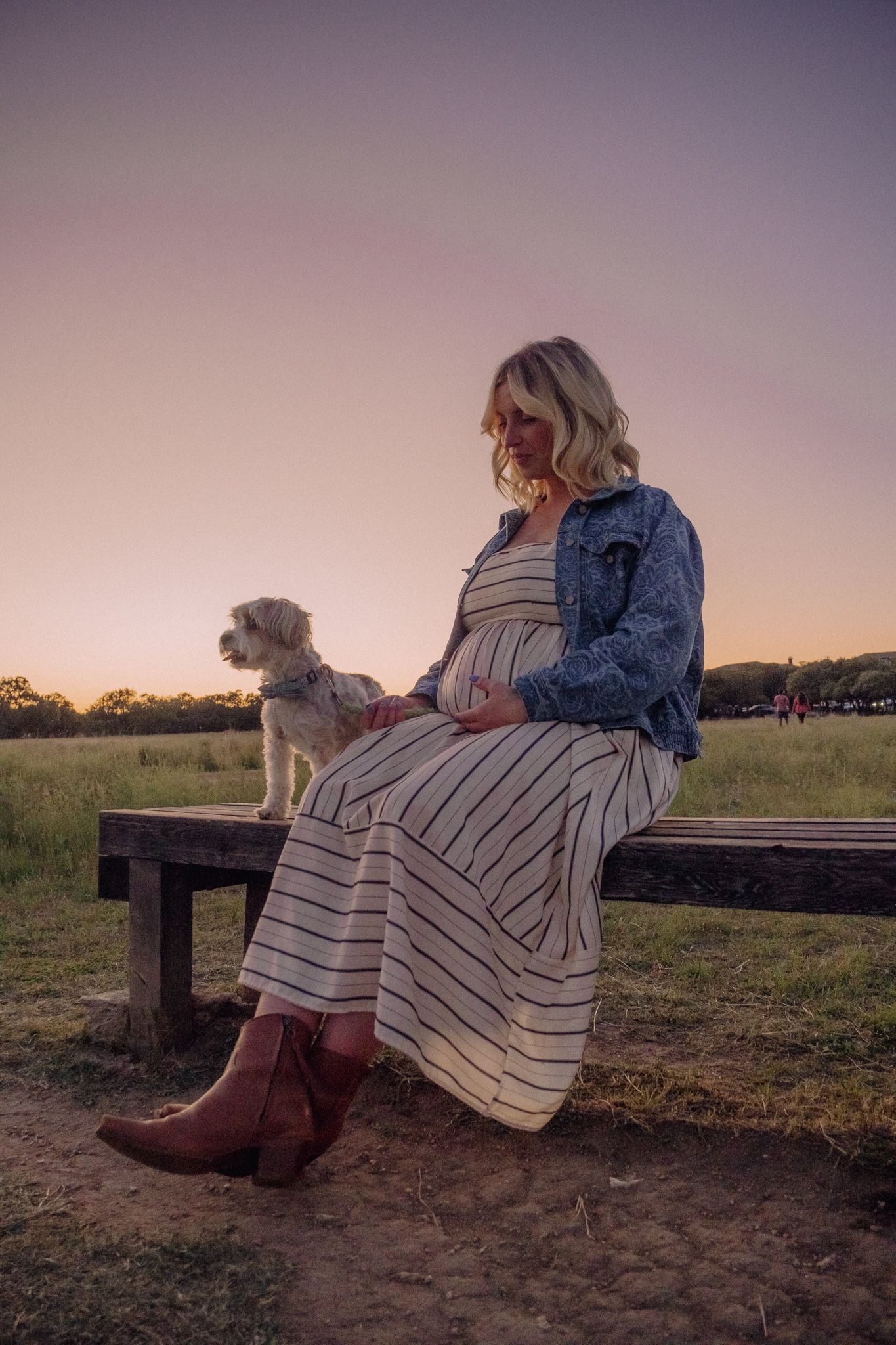 A person in a denim jacket and striped dress sits on a wooden fence at sunset with their dog in a field.