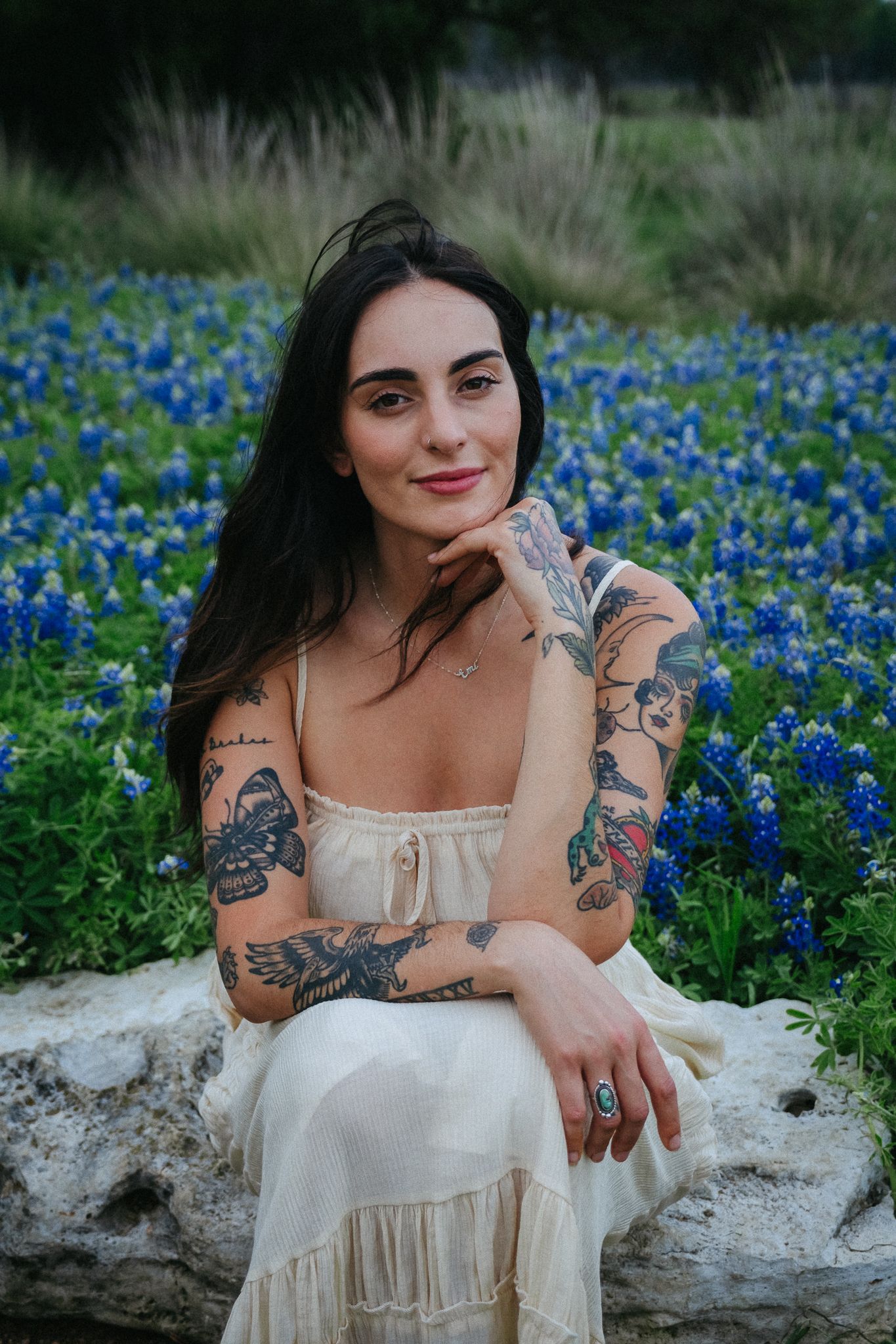 A woman in a white dress sits amid blooming bluebonnets showing her artistic arm tattoos in an outdoor portrait.