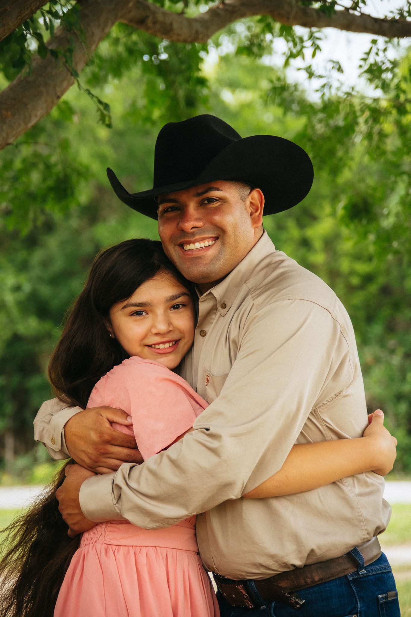 A smiling man in a black cowboy hat hugs a young girl in a pink dress outdoors near green trees.