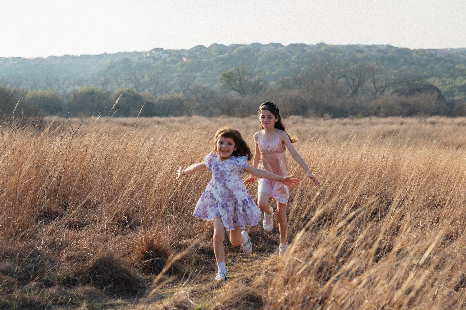 Two girls in floral dresses run joyfully through a golden field with wooded hills in the background.