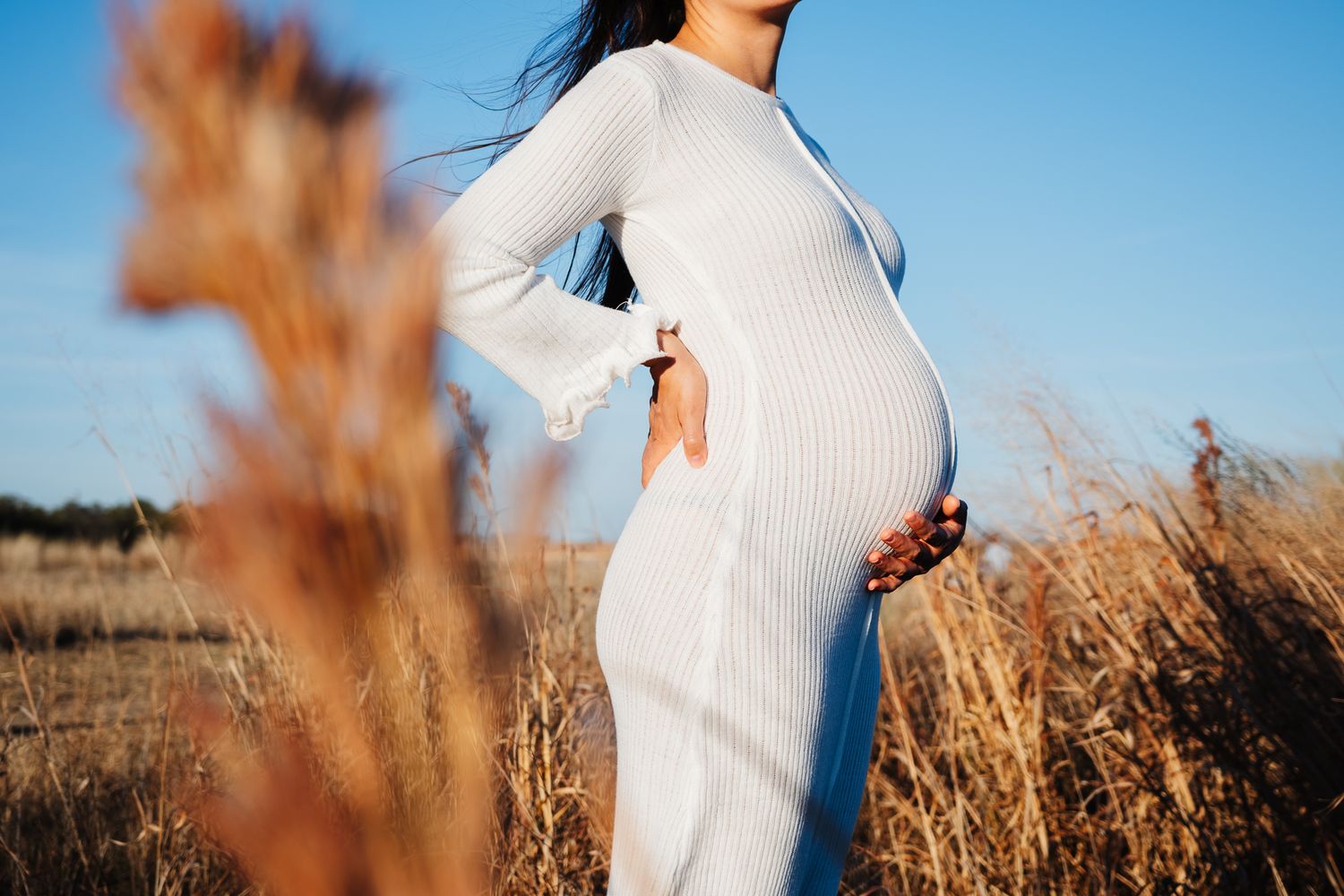 A pregnant figure in a white fitted dress poses against a backdrop of golden tall grass at sunset.