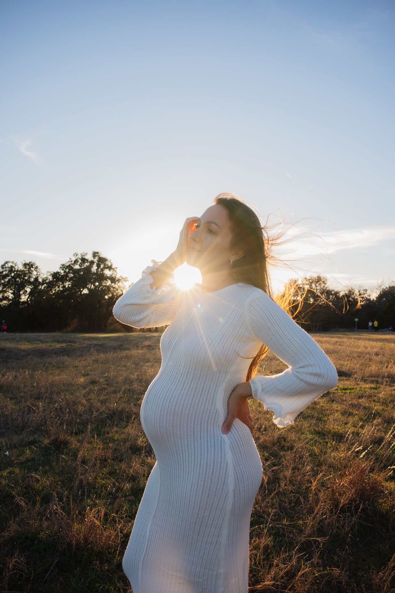 A pregnant woman in a white ribbed dress poses in a golden field at sunset as sunlight creates a warm backlit glow.