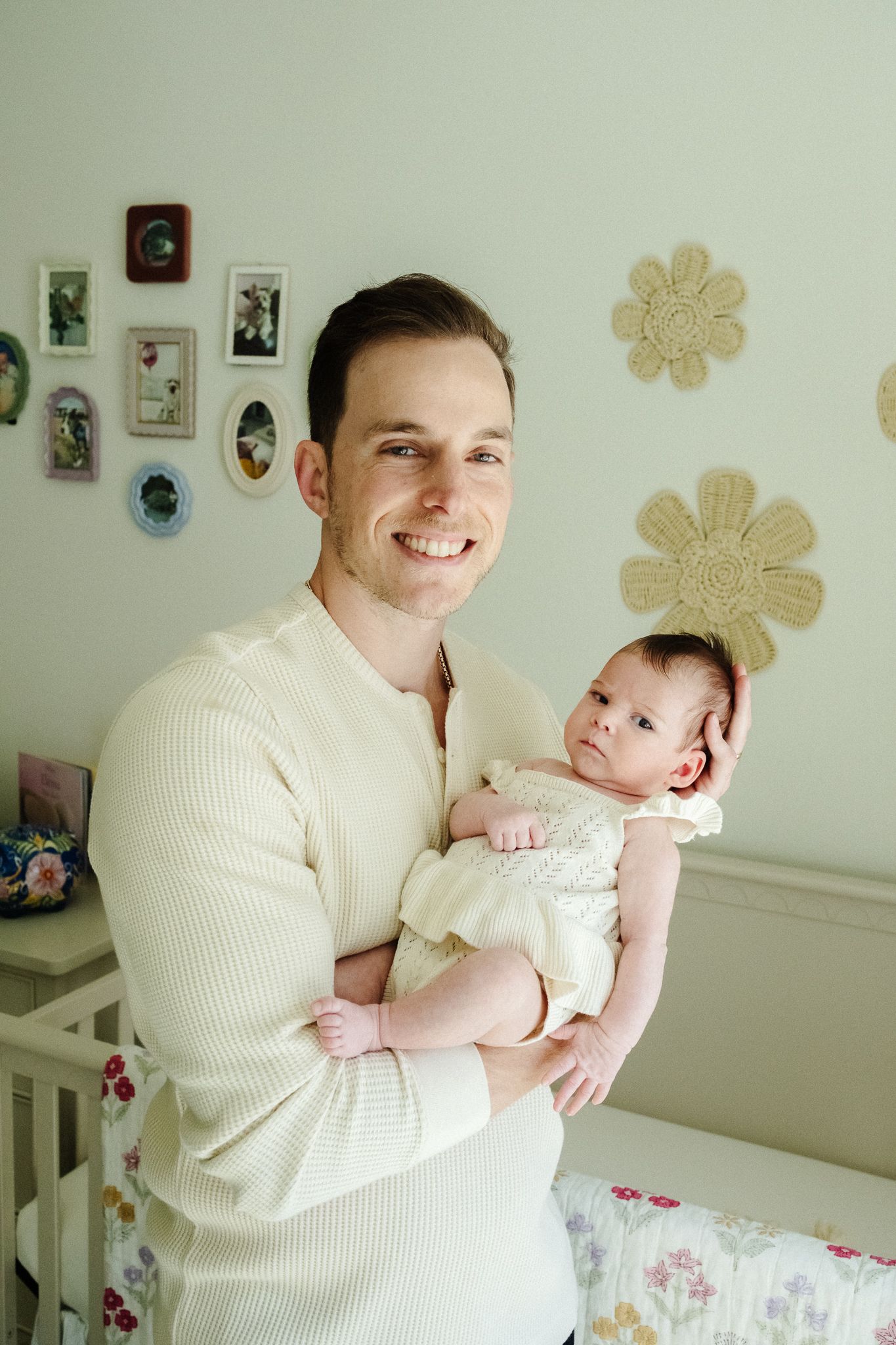 Smiling father in cream sweater holds newborn baby in decorated nursery with floral wall art and framed photos.