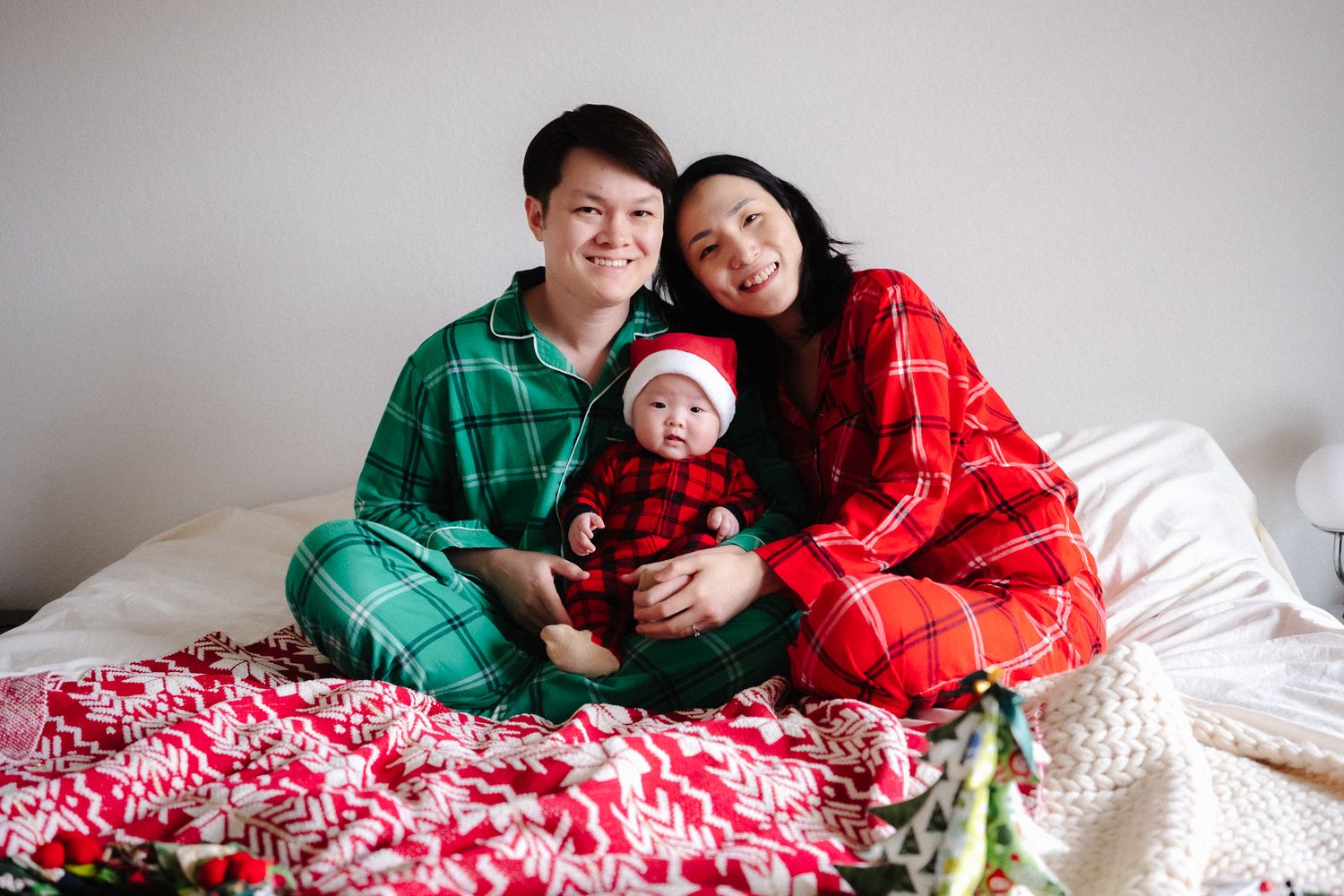 Family of three in matching Christmas pajamas sitting on a bed with holiday blankets, smiling for a festive photo.