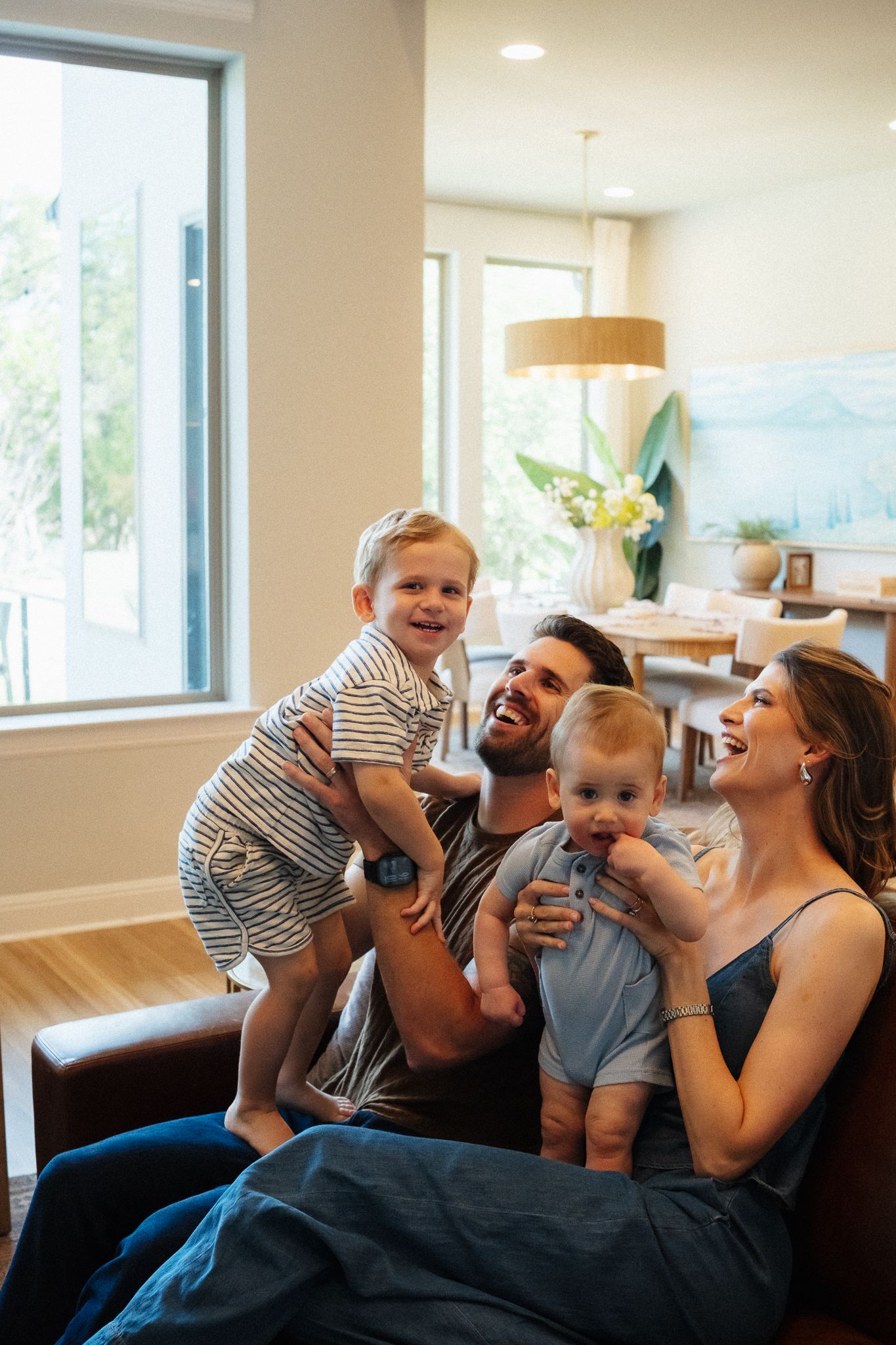 Happy family with two young children laughing together on a leather sofa in a bright modern living room.